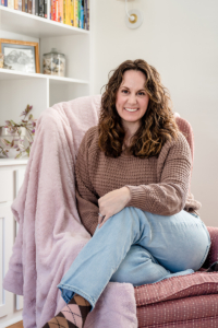 Lindsay Tiwald NJ Brand Photographer sits in a pink chair with a cozy pink blanket. Lindsay ie wearing a brown sweater and blue jeans.