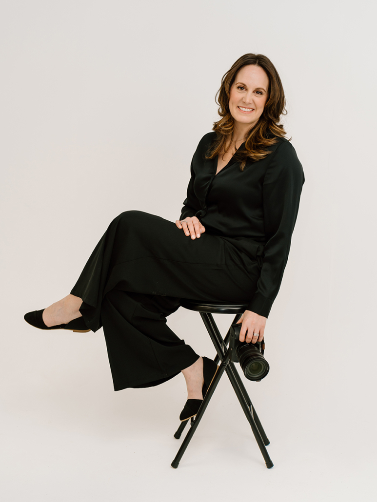 Lindsay Tiwald is dressed in al black on a white background, holds a camera while sitting on a black stool.