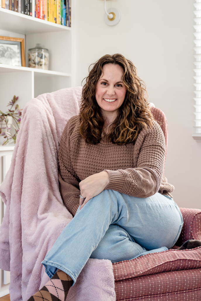 Lindsay Tiwald NJ Brand Photographer sits in a pink chair with a cozy pink blanket. Lindsay ie wearing a brown sweater and blue jeans.